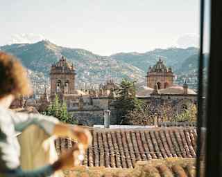 A woman in soft-focus foreground sits on the sill of an open window gazing towards Cusco Cathedral and the mountains beyond.