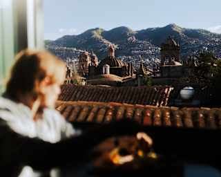 A guest in soft-focus foreground gazes through a window at Cusco's terracotta roofs and incredible mountain ranges beyond.