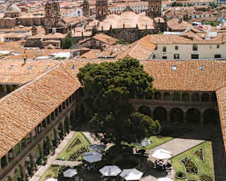 Aerial view over Monasterio's courtyard with parasols, lawn and ancient cedar tree, to Cusco's terracotta rooftops and hills.