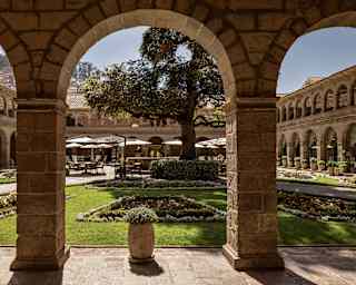 View of the courtyard with lush lawn sections, dining sets with parasols and a central cedar tree, viewed from the cloisters.