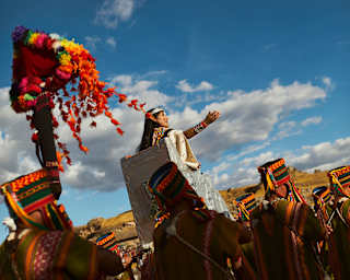 Angled view of an Inti Raymi parade with the Sapa Inca carried above the procession in a chair, arm outstretched the crowds.