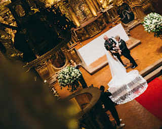 A bride and groom share a kiss at the altar of an ornate, gold-adorned church, surrounded by floral arrangements, with the scene viewed from above at an angle.