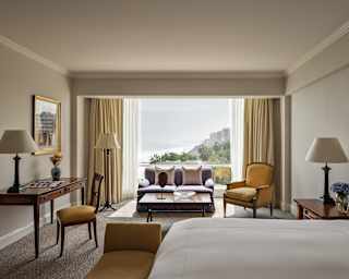 View from the bed in an Ocean View Junior Suite to the chic living area with desk, sofa, armchair and window onto the coast.