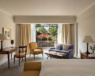 View from the bed of the Garden Junior Suite, with a desk, two yellow chairs, blue sofa, coffee table and doors to a patio.