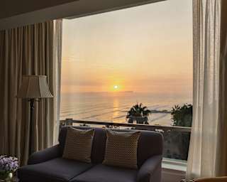 A sunset spills golden light over the waves in a view through a large window, seen across the living area of an Ocean View Suite.