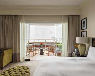 Two chairs on a balcony enjoy a misty city vista, viewed from inside a Terrace Suite over a bed and green velvet furnishings.