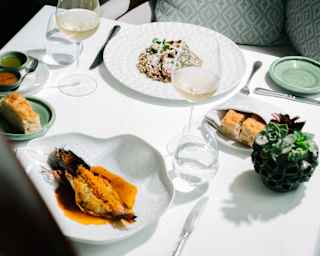 Looking down on two gourmet dishes served at an immaculate white table with a flower pot, white wine and bread, at Tragaluz.