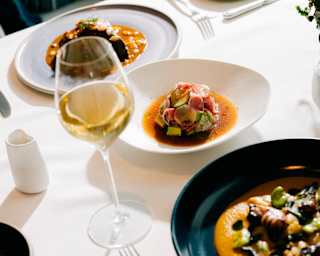 A table set with elegant crockery, including plated gourmet meals and a glass of white wine, on a white tablecloth in a bright, upmarket dining setting.