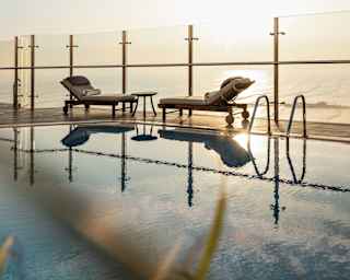Two half-reclined sun-loungers face each other over a small table by the glass terrace barrier, reflected in the still pool.