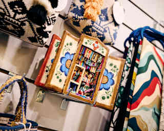 A detailed retablo, featuring a diorama of a textiles market stall and seller, hangs among bags and scarfs on a shop wall.