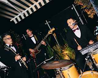 Angled image of two guitarists and a drummer, with kit, dressed in tuxedos as they play beneath a striped awning at a party.