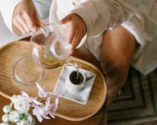 A guest in a white robe pours a cup of pale tea into a mug set on a marbled saucer, seen close-up from above at Zest Spa.