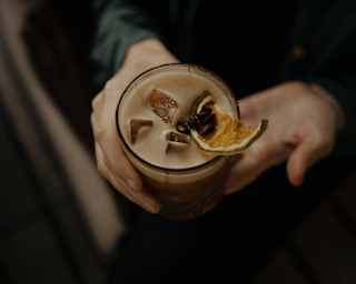 A guest holds a cocktail in a tumbler with a dried citrus slice resting on Pisco-like foam, and ice cubes, seen from above.