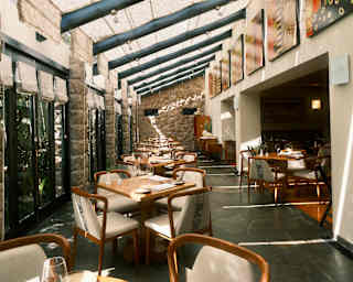 Along a wall of windows and beneath an opaque ceiling, six wooden tables with cream-seated chairs wait for the next sitting.