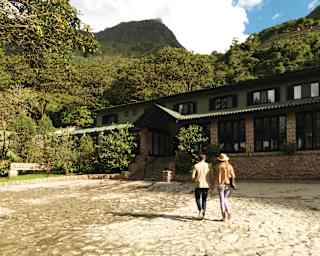 A man and woman, seen from behind, walk across a yard towards Sanctuary Lodge, nestled in forest between mountain ridges.