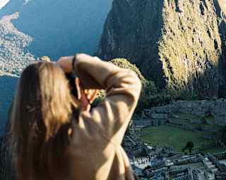 A woman with long brown hair angles her camera to get a shot of Huayna Picchu, divided by sun and shade, seen from behind.