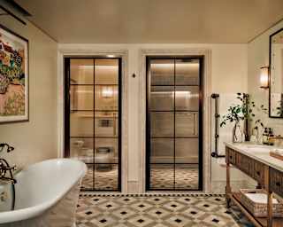 Two mirrored doors reflect geometric floor tiling in a view of a suite bathroom with bath tub, left, opposite marble basins.