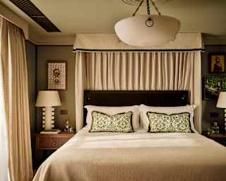 An Art Deco alabaster ceiling lamp hangs above a beige bed with green textile cushions in a view of the Artichoke Suite bedroom.