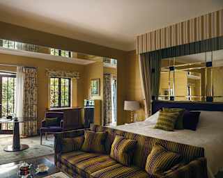 The purple of a pair of small armchairs is picked out in the amethyst stripes of a mustard sofa and pillows on the white quilt bed