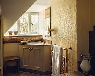 A vanity unit is tucked into the corner of wheat yellow bathroom, with brown floor tiles below an exposed oak window lintel