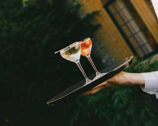 Two clear-spirit cocktails, one with fruit pieces, catch the light as they sit on a tray carried by a waiter, seen close-up at an angle.