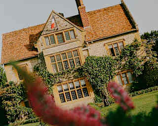 Angled view of Le Manoir in sunshine, seen across lawned garden, part-obscured by a pink lupin flower.