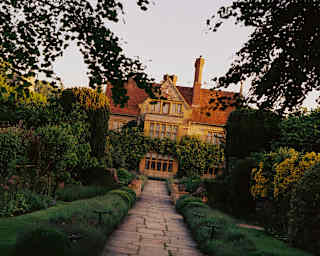 Tilted image taken on a garden path, bordered by hedges and lush gardens, leading to Le Manoir, its stone gold in late sun.