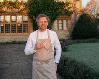 Chef Arnaud Donckele stands on a path in front of Le Manoir, smiling to camera and holding spectacles, face in late sun.