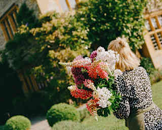A florist with blonde hair and a spotty brown dress carries a large bunch of red, white and purple lupins, seen from behind.