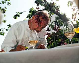 Low-angle image of Chef Raymond Blanc in spectacles as he reads at a table with two glasses of sparkling rosé in the garden.
