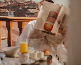 In soft-focus foreground, a guest reads at a breakfast table on a balcony with city views, face hidden by an open newspaper.