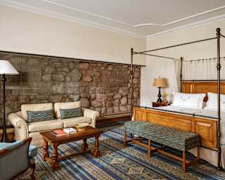 View over a woven blue rug in an Inca Suite to an exposed stone wall by a lounge area with a pale sofa and a four-poster bed.