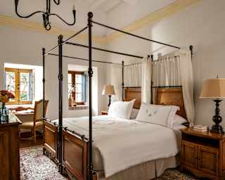 Diagonal view over a Junior Suite room with a four-poster bed and side tables to a wall with two window alcoves and a desk.