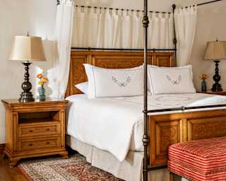 View of a four-poster bed in a One Bedroom Suite with white drapes gathered behind the walnut headboard, and a side table.