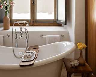 A wooden tray with two rolled flannels spans a slipper bath by a light-filled window in a bathroom with white and wood décor.