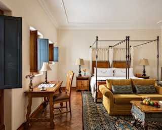 A wooden desk and chair bathes in light from a window above, in a view through a Studio Suite with coffee table, sofa and bed.
