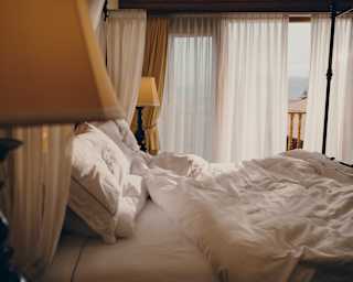 Morning light drifts through translucent drapes drawn across glazed doors, seen past a side lamp and over a rumpled white bed.