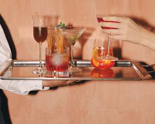 A guest's hand picks a cocktail from a choice of six, in a variety of glasses and hues, from a silver tray held by a waiter.