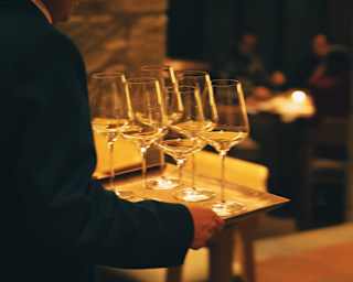 A waiter carries five elegant wine glasses on a tray through the warm-lit restaurant, seen in close-up from the side.
