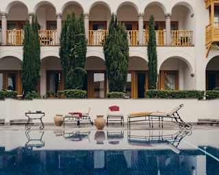 View of three loungers with white cushions and yellow and red towels, overlooked by balconies, seen over still pool waters.