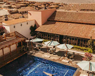 A guest floats in the azure pool, lined with parasols in a courtyard of terracotta-tiled buildings, seen from above.