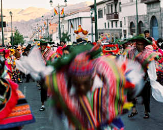People in traditional brightly-woven shawls parade through a street with rainbow flags on lampposts, blurred with movement.
