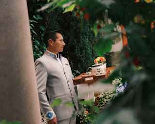 A smart male member of staff stands in a vine-covered courtyard holding a wooden tray with a teapot, seen through foliage.
