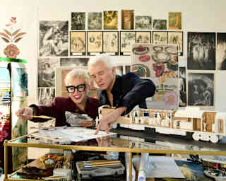 Baz Luhrmann, in black, and Catherine Martin in burgundy with glasses, look through designs in a busy art studio.