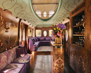 Luxurious vintage train carriage interior with ornate floral decor, plush purple seating, an oval ceiling mirror, flowers on a gold stand, and a built-in bar, all bathed in warm, elegant lighting.