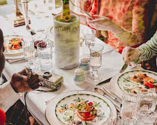 Four guests enjoy a vibrant lunch at a table busy with glasses of red wine, water and Champagne, seen from above, close-up.