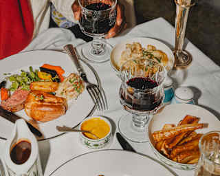 A diner is glimpsed holding a glass of red wine at a table with two plated roast beef dinners with vegetables and gravy.