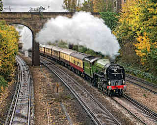 A plume of steam billows out along the length of the cream and burgundy carriages as the train passes below an arched bridge