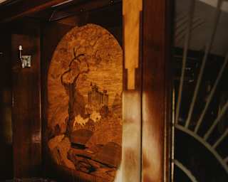 A wooden wall panel featuring an engraved tree and landscape scene is next to a sofa with patterned upholstery and a window with decorative glass. Warm sunlight highlights the cosy, vintage interior.