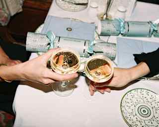 A couple chinks coupe glasses of fizzing Champagne over a table laid with Christmas crackers, seen close-up from above.